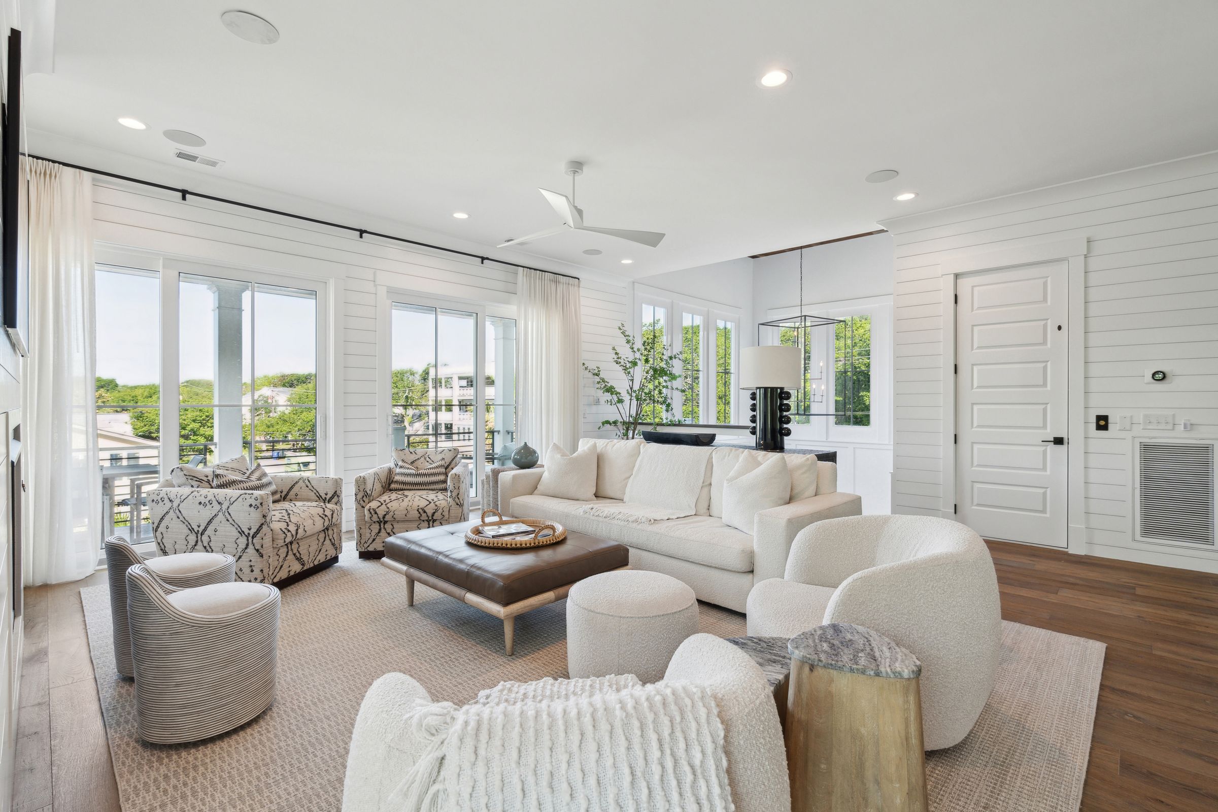Bright, modern coastal living room with white shiplap walls, neutral-toned furniture, sliding glass doors, and natural light overlooking a balcony view.
