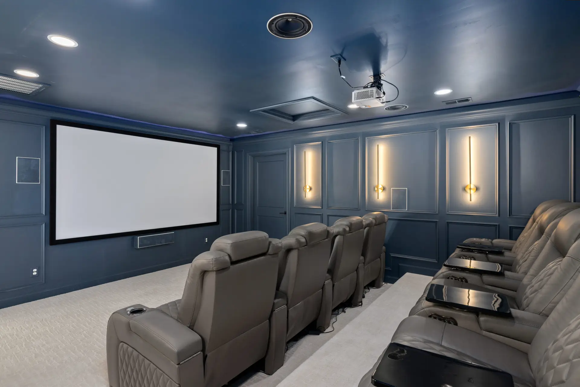 Luxury home theater with navy blue walls, leather recliner seating, wall-mounted lights, ceiling projector, and a large screen in a dedicated media room.