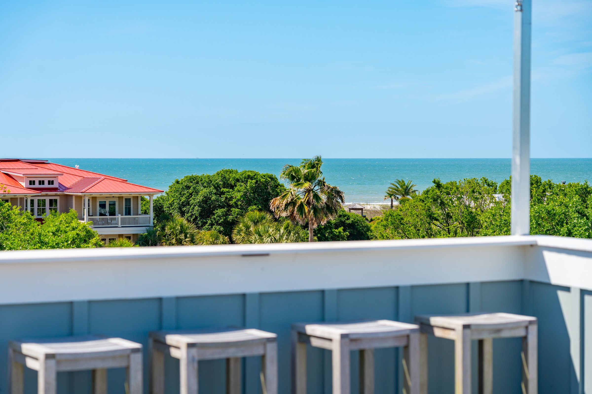 Ocean view from a rooftop deck with bar stools overlooking palm trees, a red-roofed beach house, and the Atlantic coastline on a clear, sunny day.