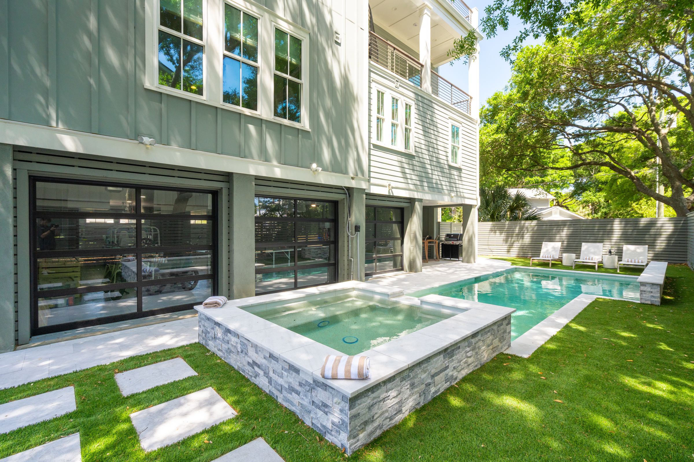 Modern backyard with private pool and elevated spa, surrounded by lush landscaping, sun loungers, and a shaded patio area beneath a coastal-style home.